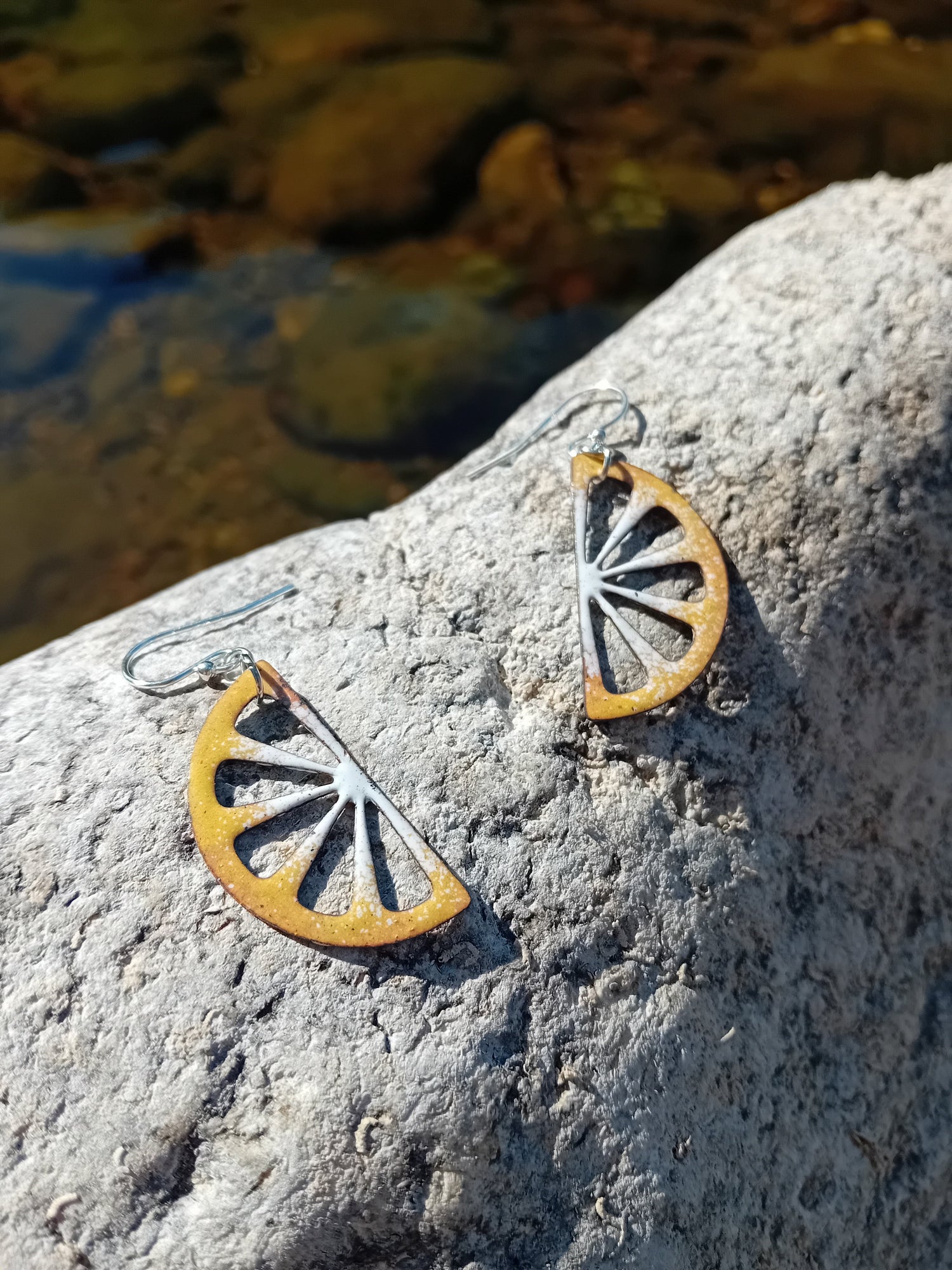 Boucles d'oreilles forme quartier de citron avec émail coloré, dégradé jaune et blanc. Les attaches sont des crochets argent, et la paire de boucles est posée sur un gros caillou au bord d'une petite rivière.